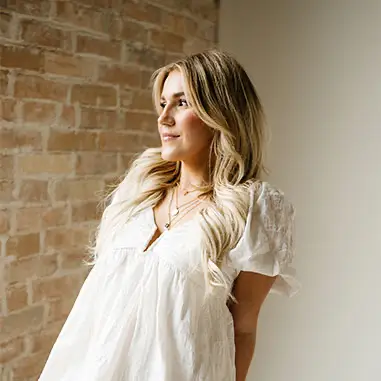 White-dressed woman poses against brick wall, endorsing Hair Conditioning Treatments.