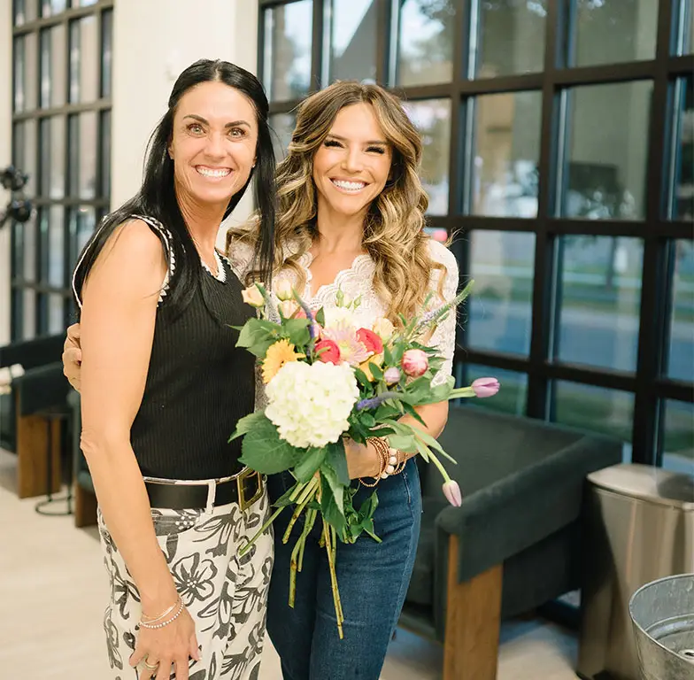 Two women smiling and holding flowers, standing side by side at Salon M + Suites. Two women smiling and holding flowers, standing side by side at Salon M + Suites.