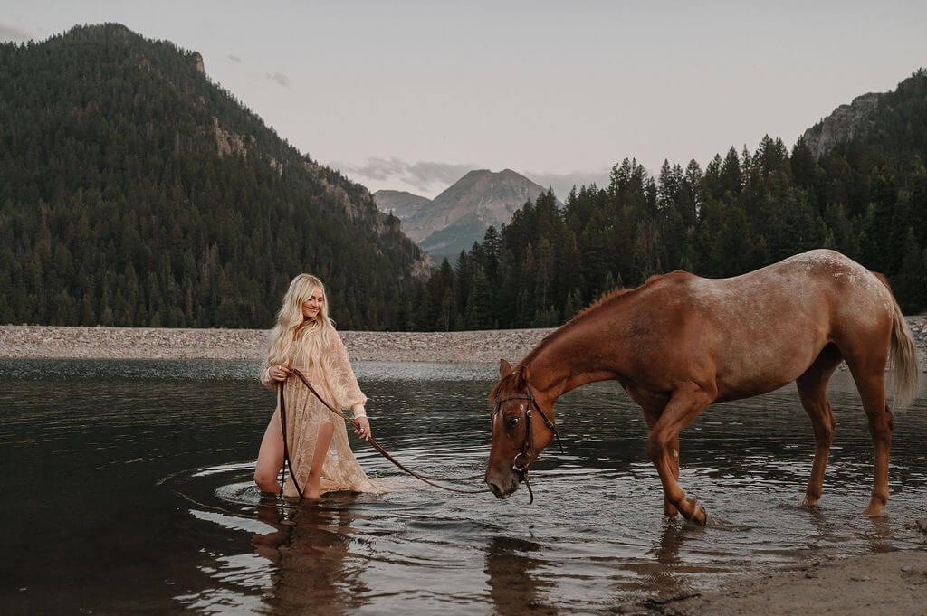 Blonde hair girl walking with horse thorugh lake in the mountians